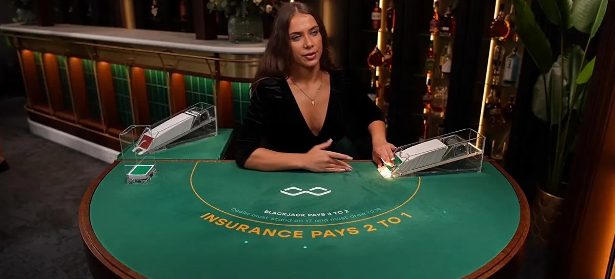 Smiling live blackjack dealer in a black dress seated at a green felt table with card shoes, chip tray, and “Insurance Pays 2 to 1” text, set in a stylish bar-like casino studio.