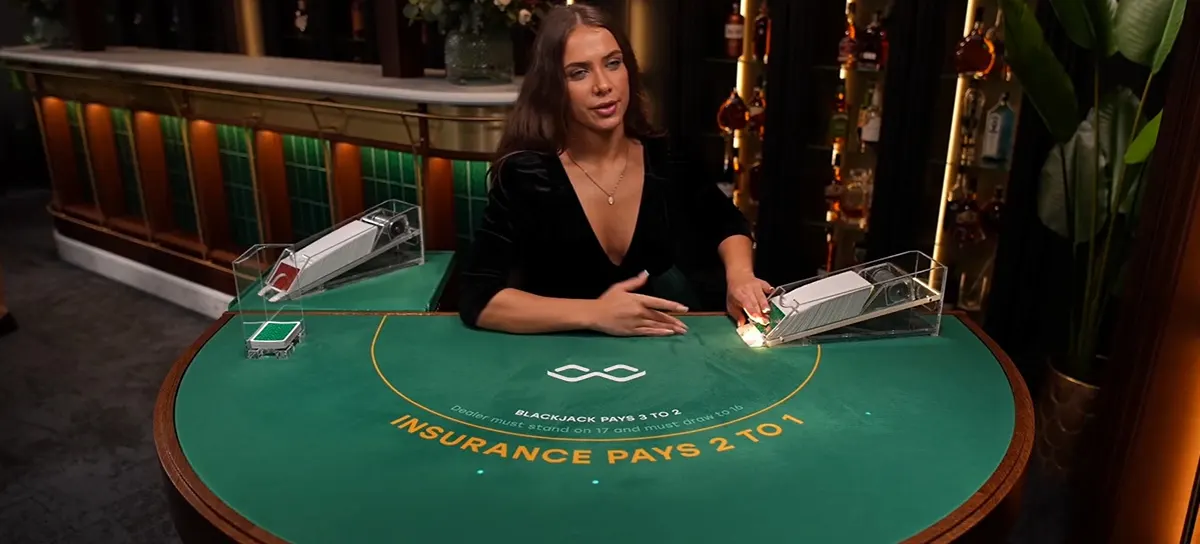 Smiling live blackjack dealer in a black dress seated at a green felt table with card shoes, chip tray, and “Insurance Pays 2 to 1” text, set in a stylish bar-like casino studio.