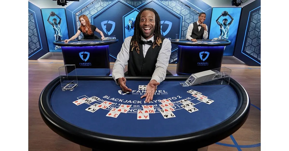 Excited live blackjack dealer in a blue FanDuel Casino studio leans over a round table with scattered playing cards, while two other dealers work at tables in the background under bright stage lighting.