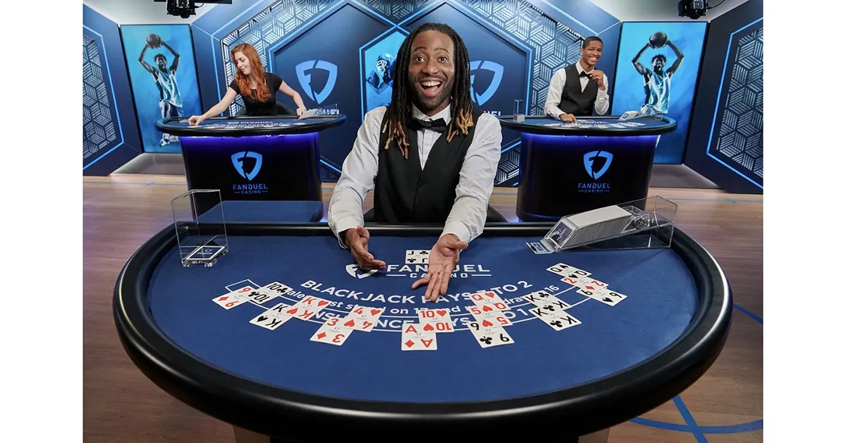 Excited live blackjack dealer in a blue FanDuel Casino studio leans over a round table with scattered playing cards, while two other dealers work at tables in the background under bright stage lighting.